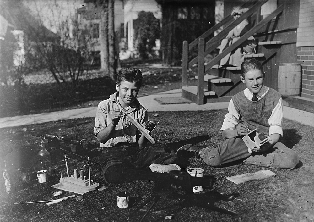 A black and white photograph of two young boys in a backyard. One is building a small wooden boat, the other painting a wooden ambulance.