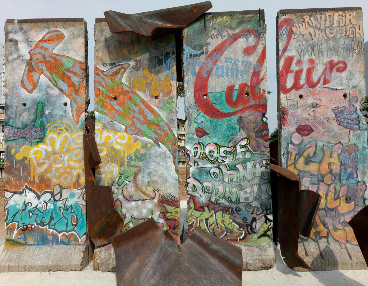 Four segments of the Berlin Wall covered in colourful paintings and adorned with metal figures, climbing over the wall and slipping through the spaces between the segments.