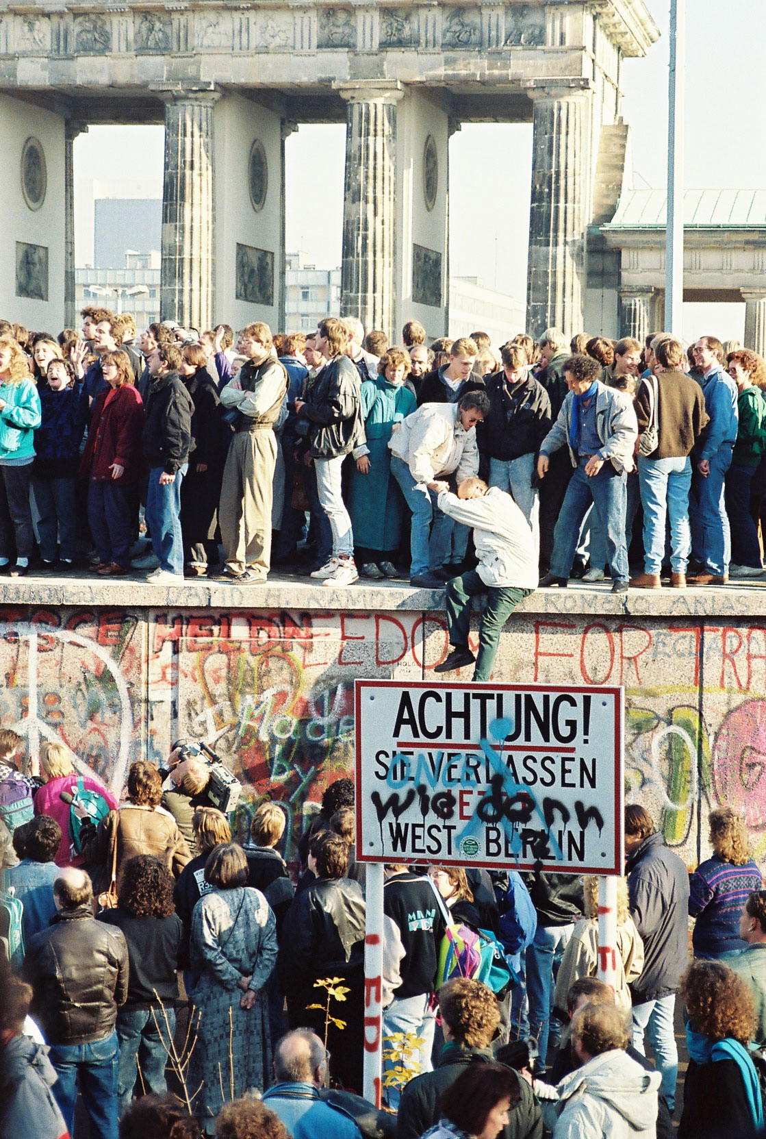 People atop the Berlin Wall near the Brandenburg Gate on 9 November 1989. A street sign reads "Achtung! Sie verlassen jetzt West-Berlin" ("Notice! You are now leaving West Berlin") has been modified with an additional text "Wie denn?" ("How?").