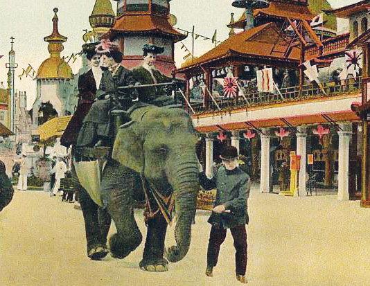 An image from a colorized postcard, showing three women riding on the back of an Elephant, being led by a young man. They are passing by a palatial structure decorated with flags.