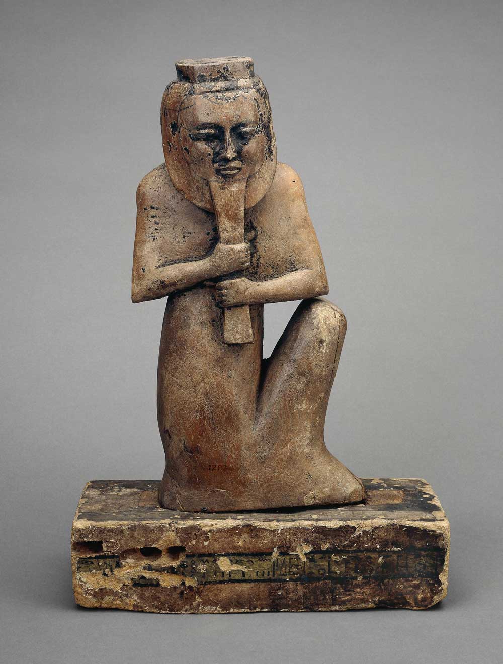 Wooden figure of a squatting deity with head turned and grasping beard in both hands.