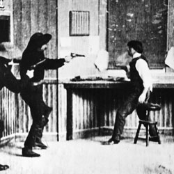 A still from a black and white film showing two people with guns drawn on a third man.