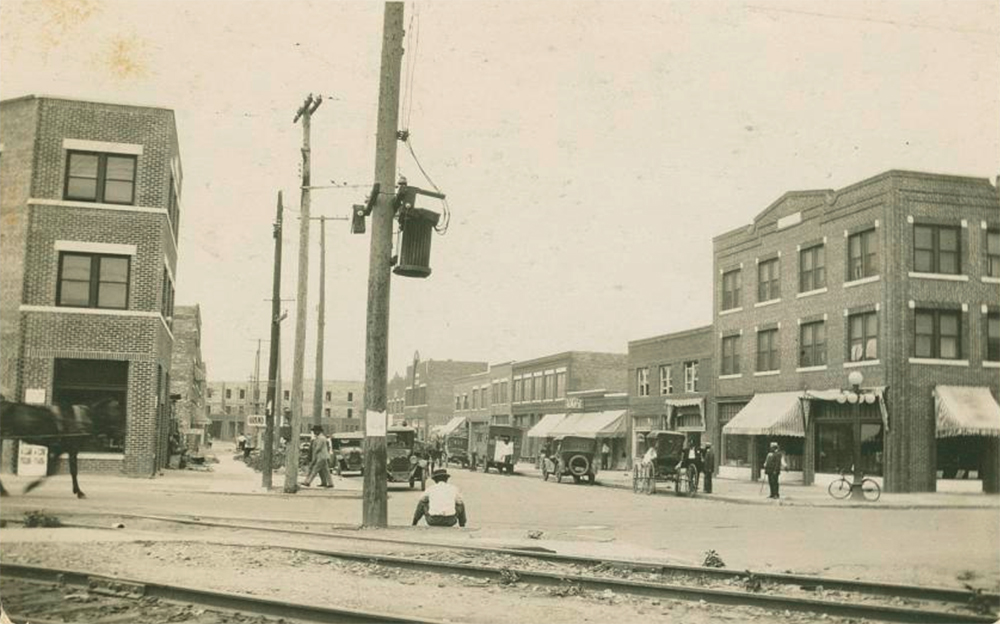 A black and white photograph of the Greenwood district in Tulsa, OK. Depicted are cars parked on a street, a horse, a carriage, people walking along shopfronts, and telephone poles.