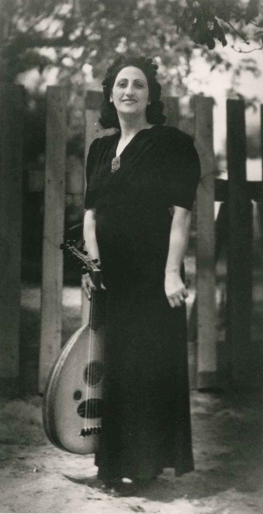 An Armenian woman in a long black dress stands in front of a fence, holding an oud.