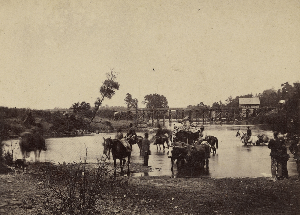 Black men on horseback fording a river with an oxcart.