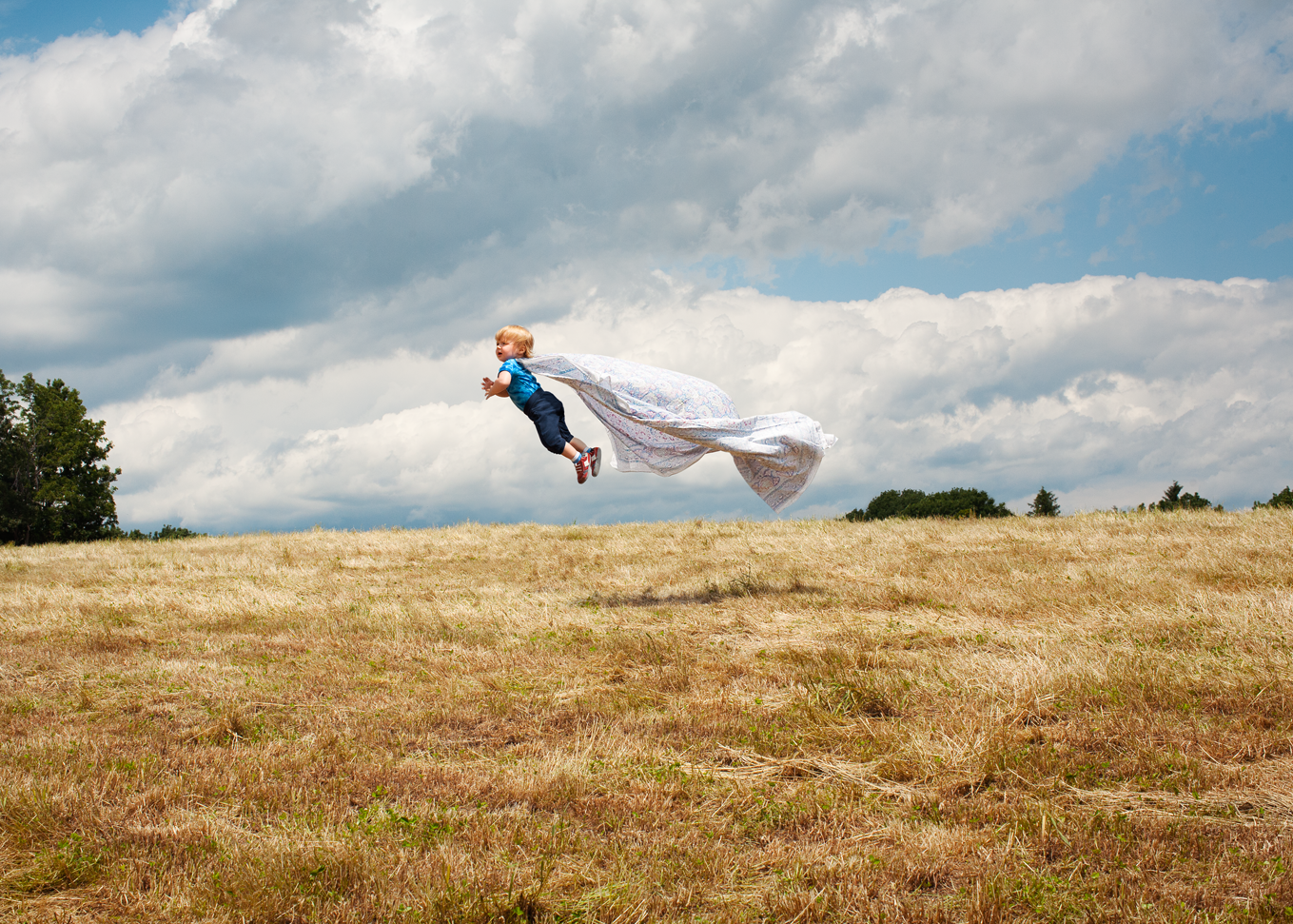 Color photograph of a toddler wearing a cape and seemingly flying in a wheat field.