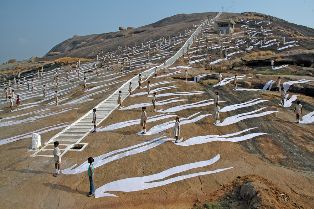 Color photograph of children standing on a hill with long white shadows trailing behind them.