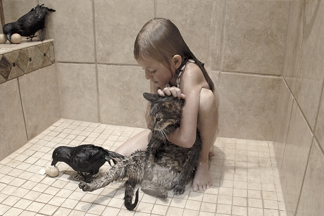 Color photograph of a young girl sitting in a shower with a wet cat and several crows.