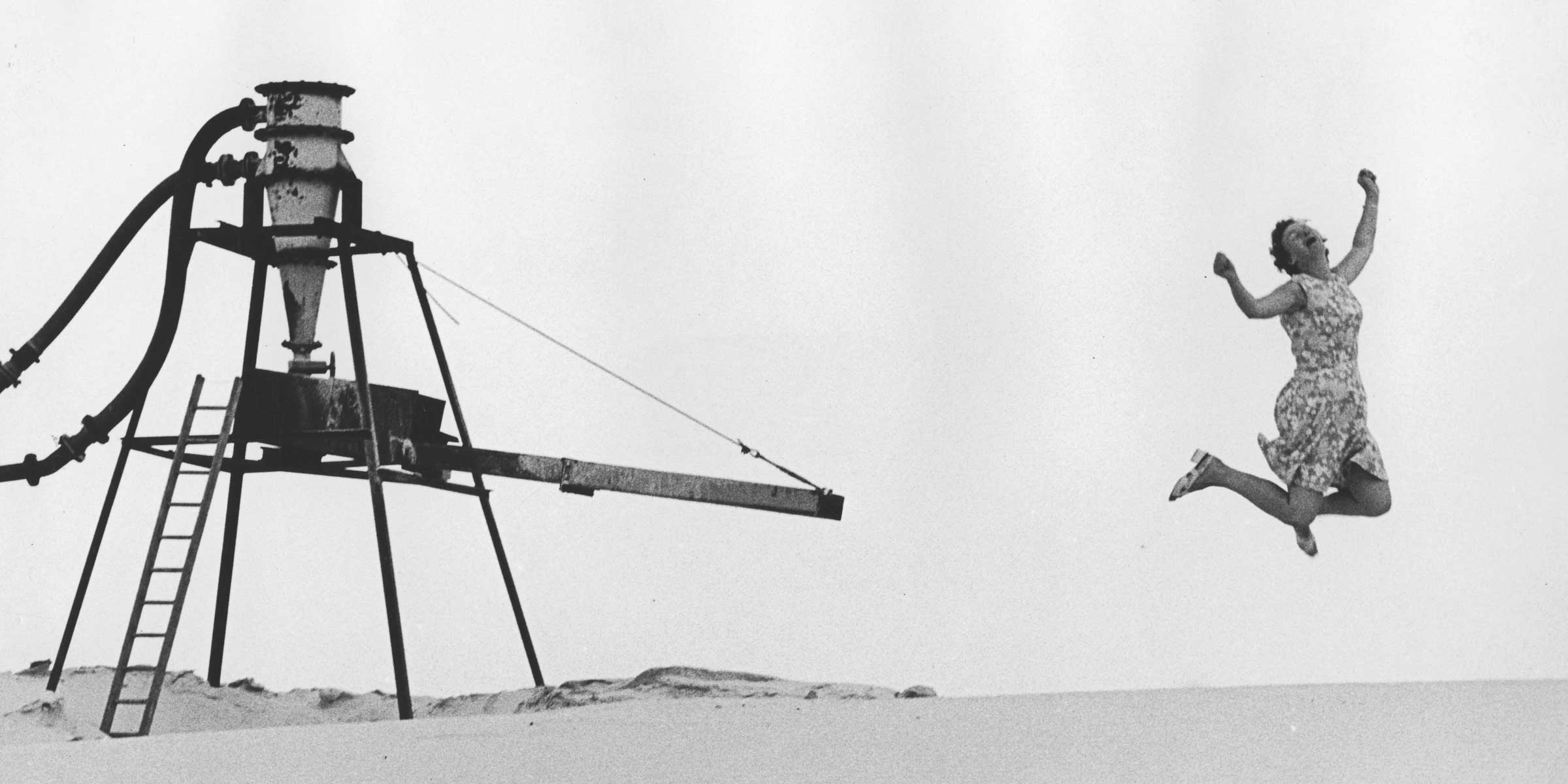 Black and white photograph of a woman jumping on a beach with industrial equipment in the background.