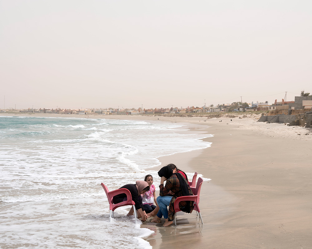 Refugees temporarily housed at a seaside resort, Castelverde, Libya, 2019. Photograph by Lorenzo Meloni. © Lorenzo Meloni / Magnum Photos. Three fully dressed children and a woman sitting in chairs partly in the water on an empty beach