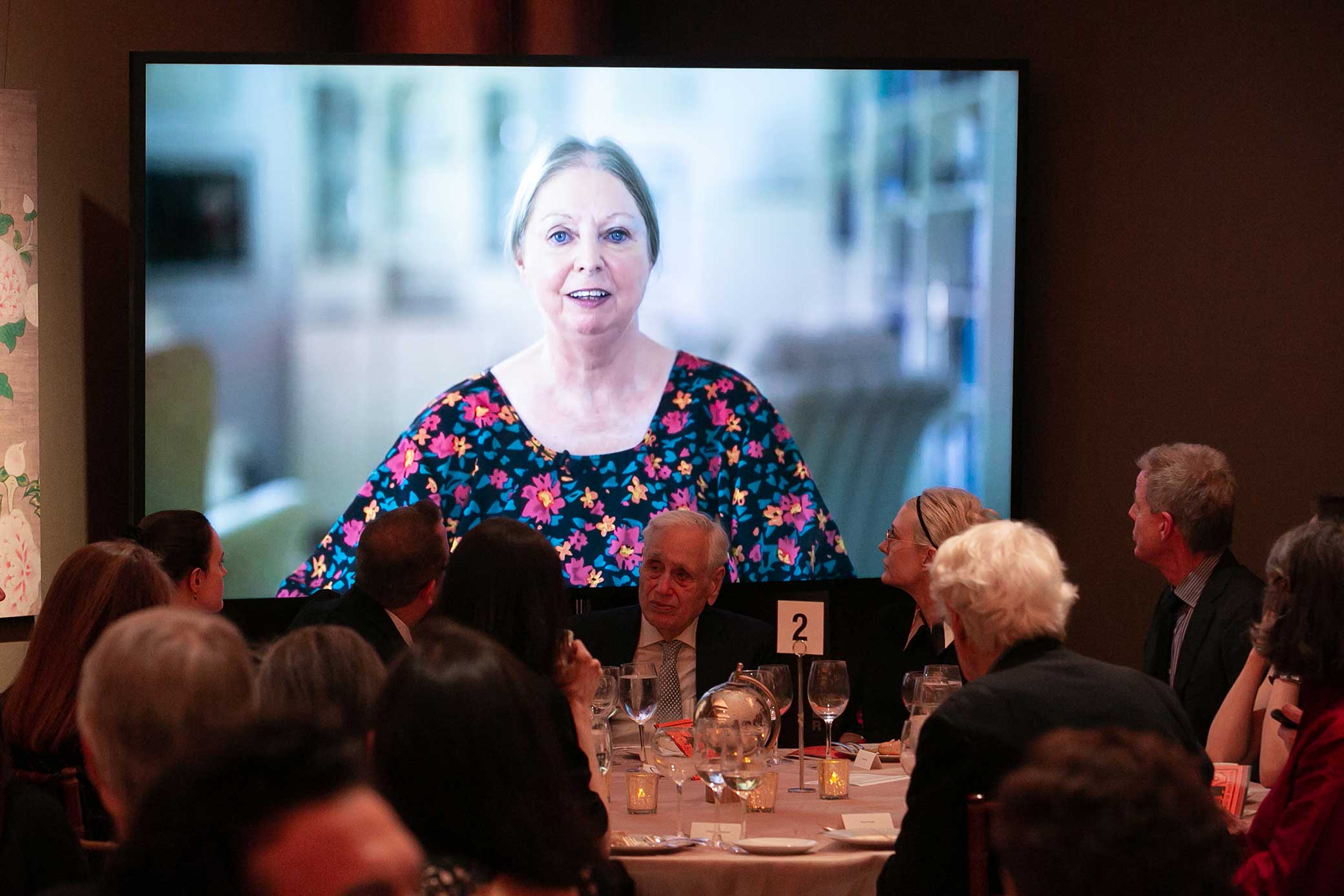 A woman in a flowered dress speaks in a a video screened behind a dinner table 