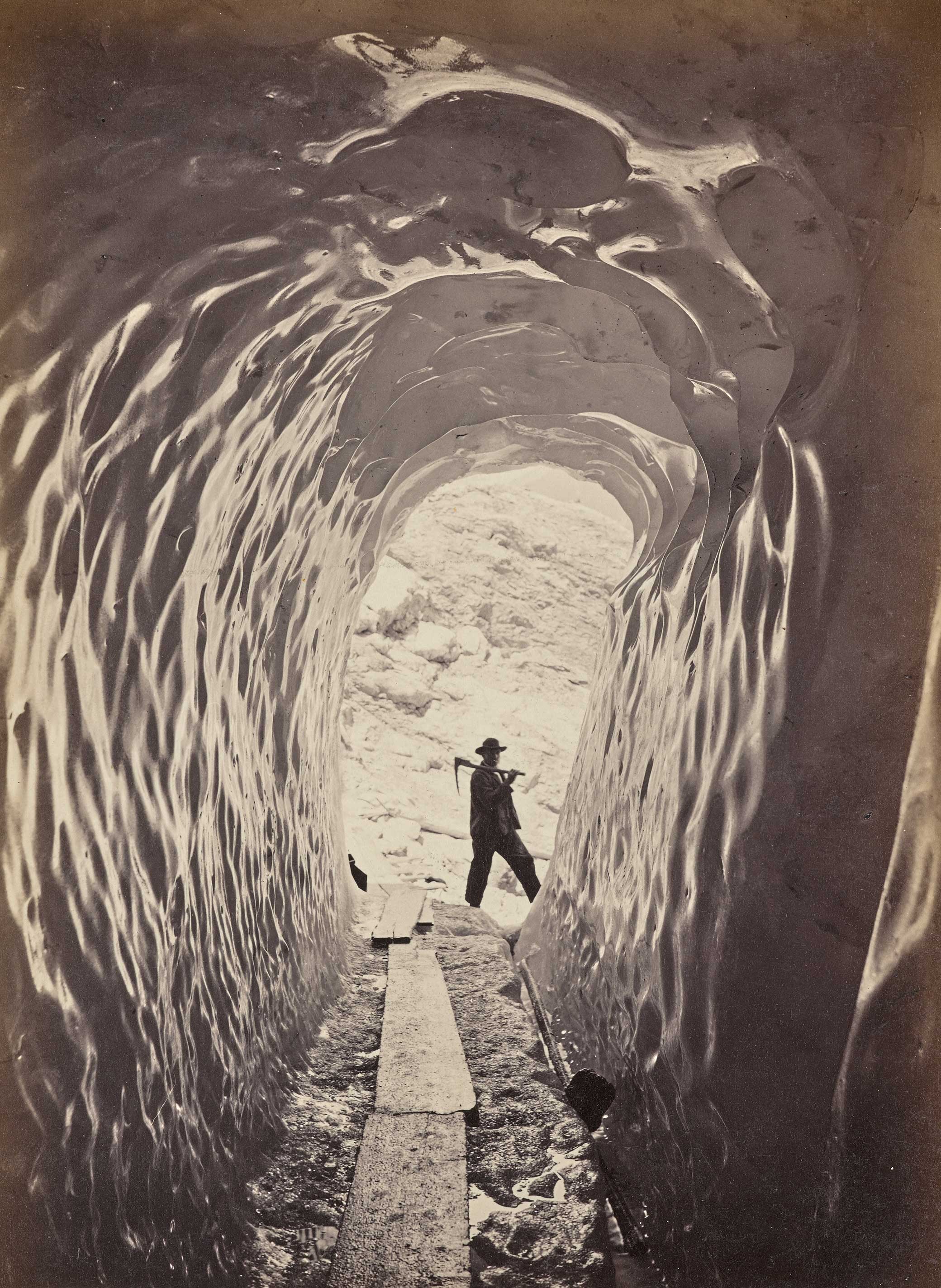 A man seen standing at the end of an ice tunnel. He has an ice pick resting over one shoulder and is turned to look down the tunnel toward the camera. Wooden planks are placed along the floor of the tunnel creating a walkway.