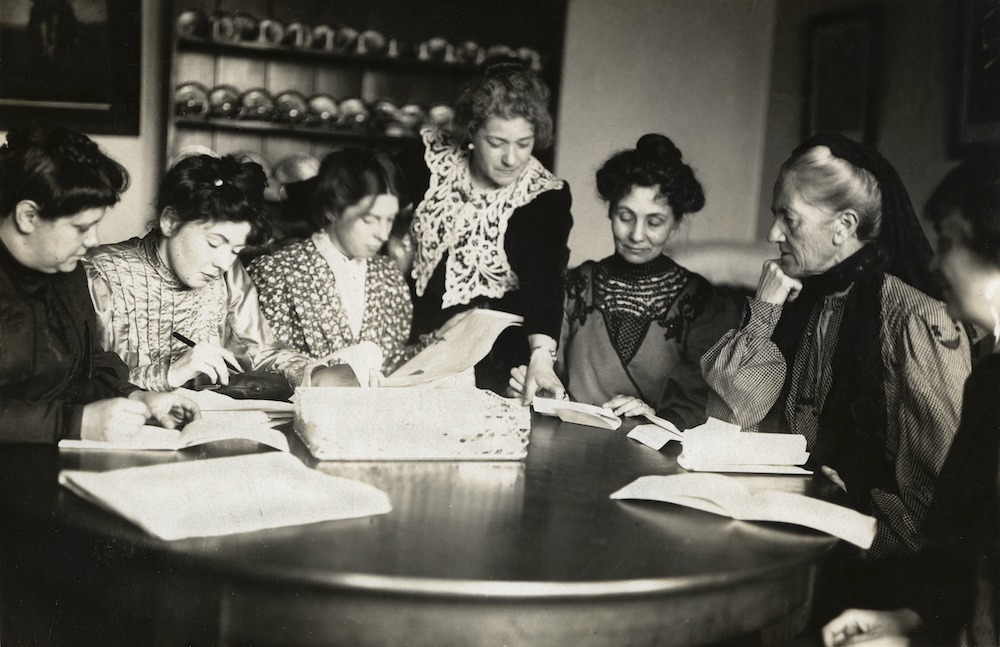 Black and white photograph of Flora Drummond, Christabel Pankhurst, Jessie Kenney, Nellie Martel, Emmeline Pankhurst, and Charlotte Despard working around a kitchen table.