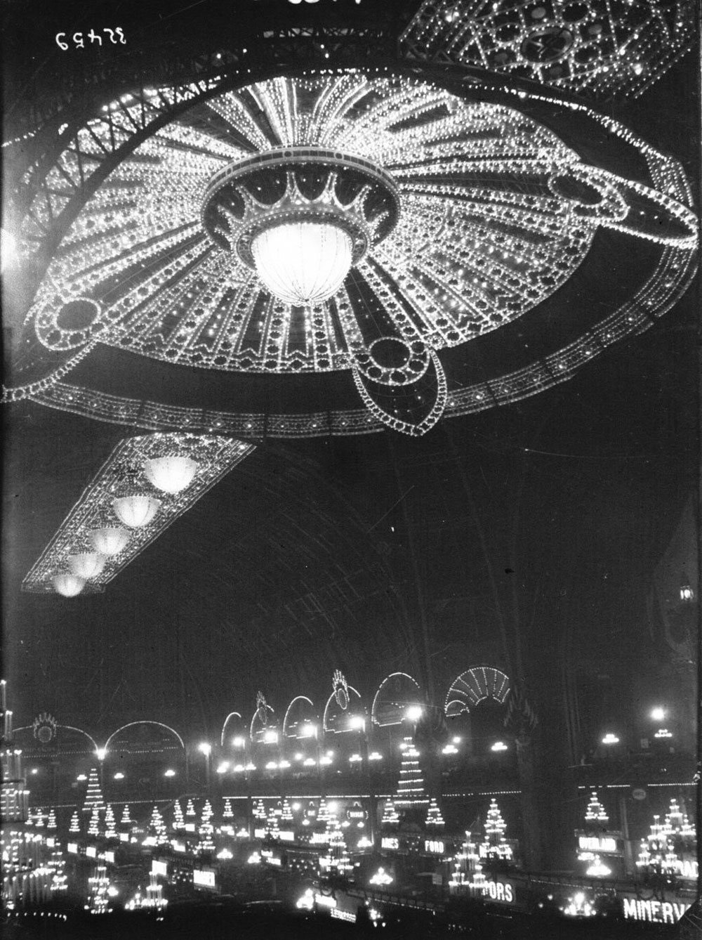 A black-and-white photograph of a large room with high ceilings. The ceiling is decorated with an ornate lighting design.