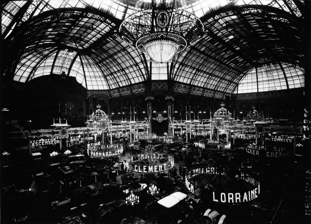 A black-and-white photograph of a large room with a domed glass ceiling. The room is full of cars, and lights above the cars spell out the names of car manufacturers.