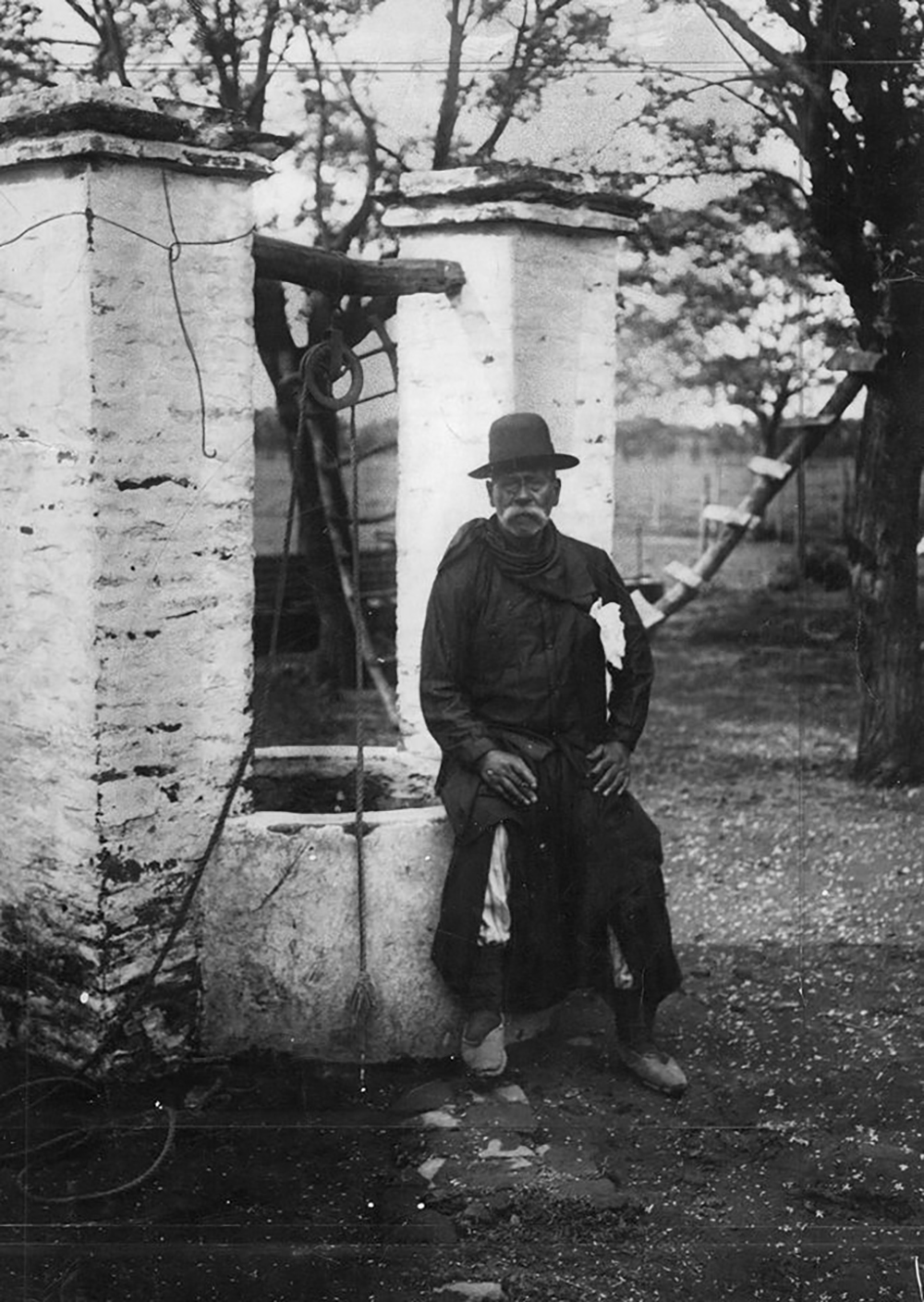 Black-and-white photograph of a man with a white mustache and hat seated on the edge of a well.