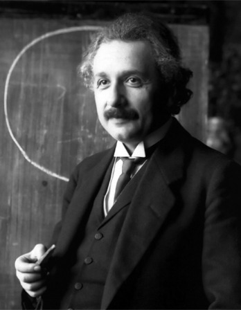 Albert Einstein in a suit and tie standing in front of a chalkboard 