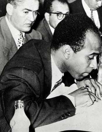 Profile of a man in a suit jacket leaning over a piece of paper with men in the background