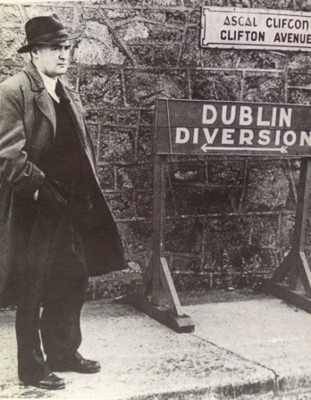 Flann O'Brien in an overcoat standing next to a Dublin Diversion sign