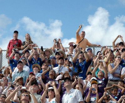 Spectators watch the launch of the space shuttle Discovery, 2007. NASA/KSC. Photograph of a crowd of people seated in bleachers and looking up.