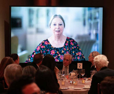 A woman in a flowered dress speaks in a a video screened behind a dinner table