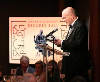 Man in a tuxedo reading at a podium with a screen in the background