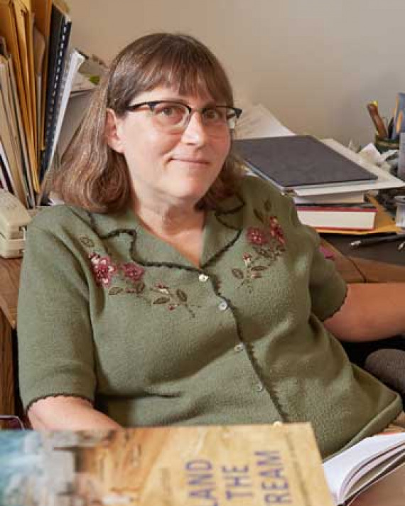 Woman wearing glasses seated at a desk with books stacked in front of her