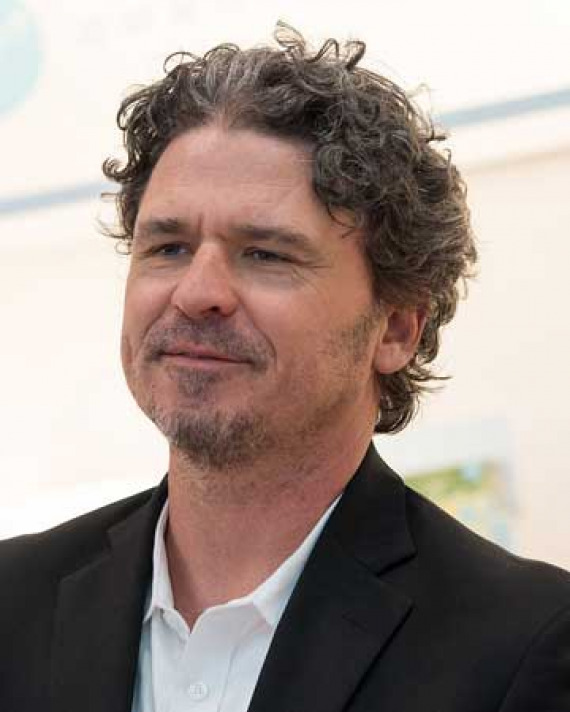 Dave Eggers Profile of a man with curly hair and goatee wearing a black jacket and white shirt