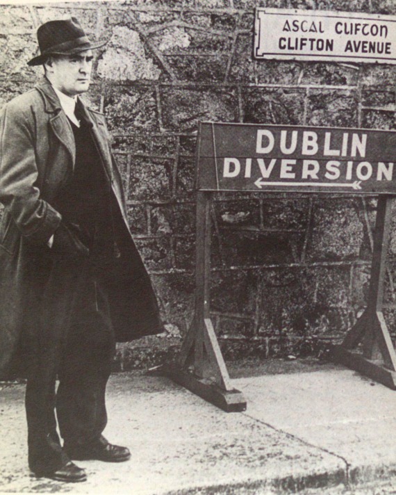 Flann O'Brien in an overcoat standing next to a Dublin Diversion sign