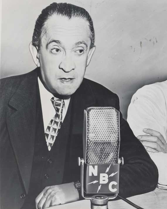 Seated man with slicked back hair speaking into a microphone