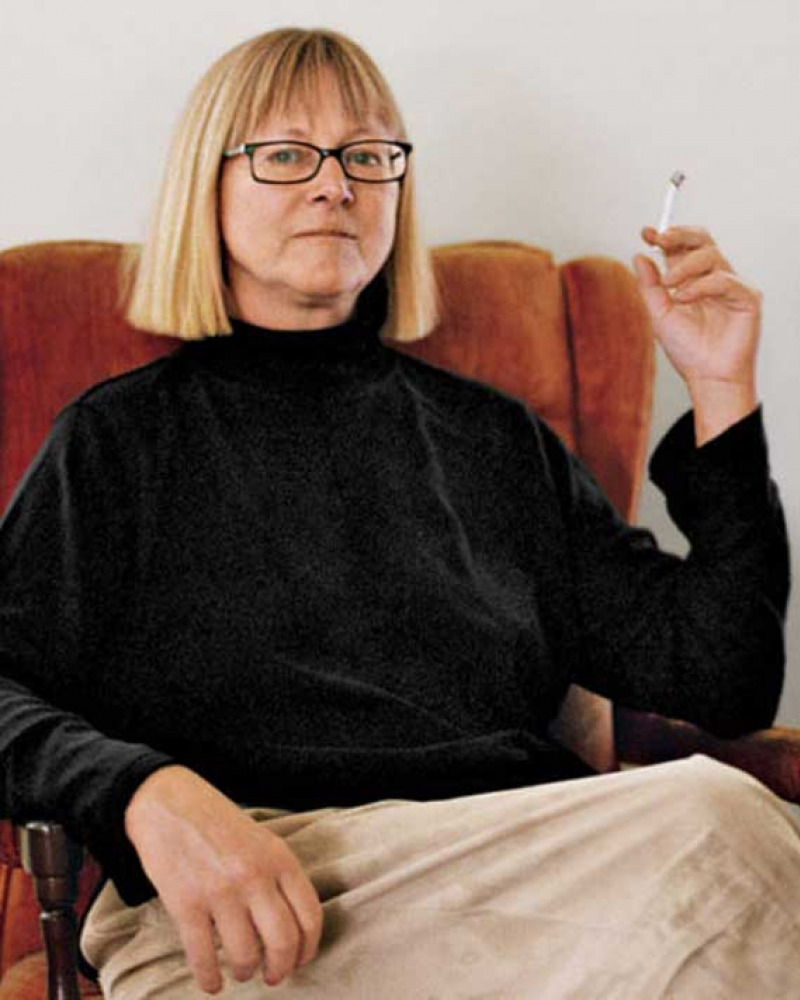 Seated woman wearing glasses and holding up a cigarette