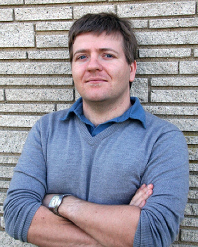 Photograph of Colin Dickey standing in front of a brick wall.