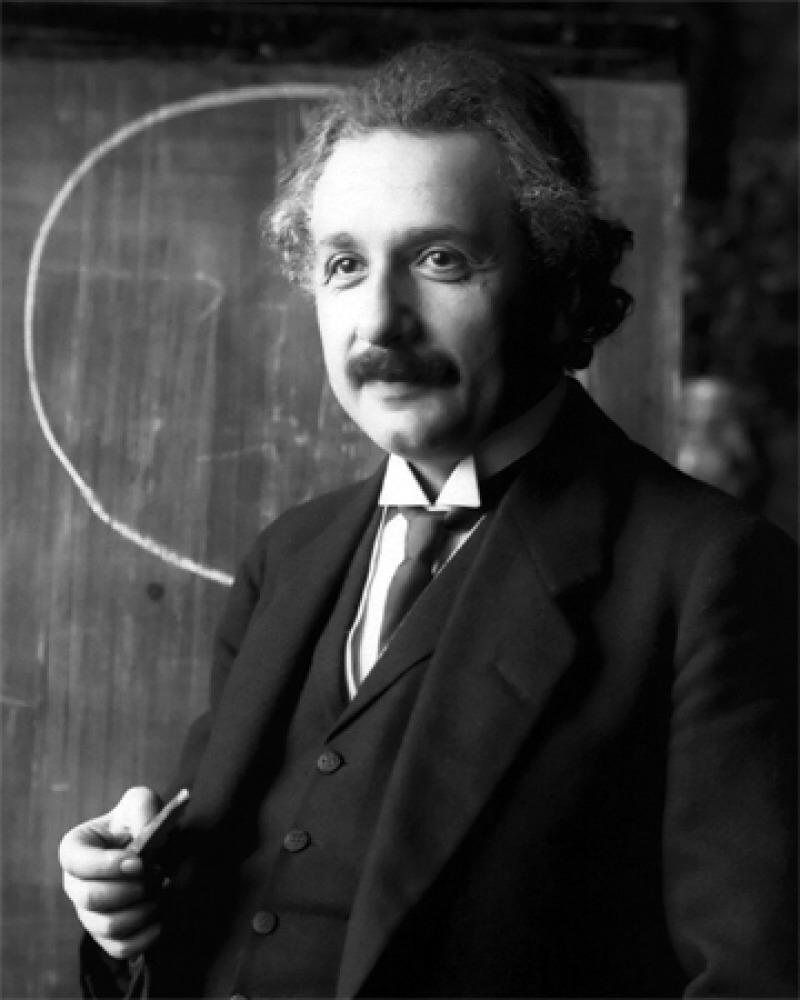 Albert Einstein in a suit and tie standing in front of a chalkboard 