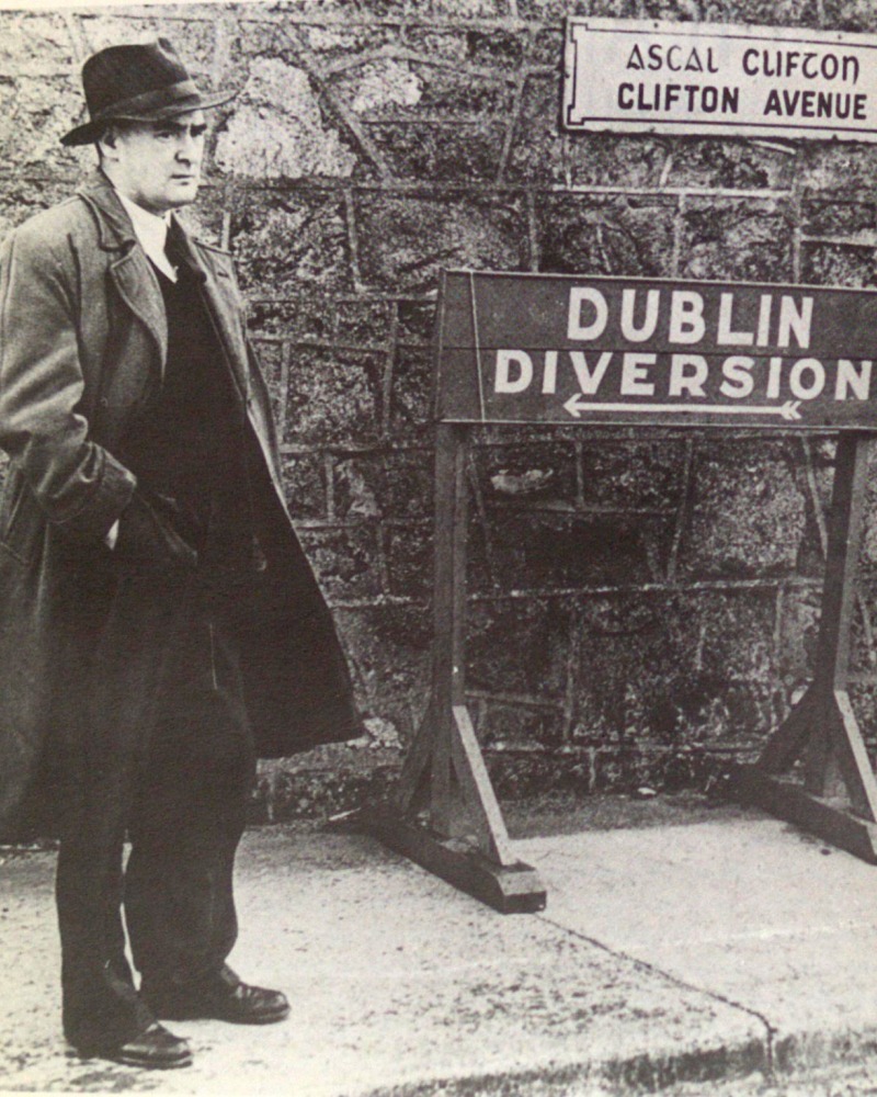 Flann O'Brien in an overcoat standing next to a Dublin Diversion sign