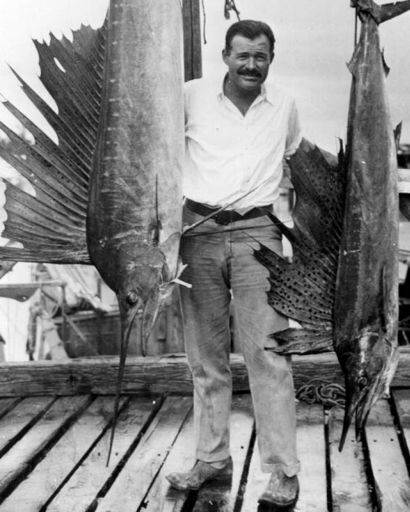 Ernest Hemingway standing between two large hanging fish