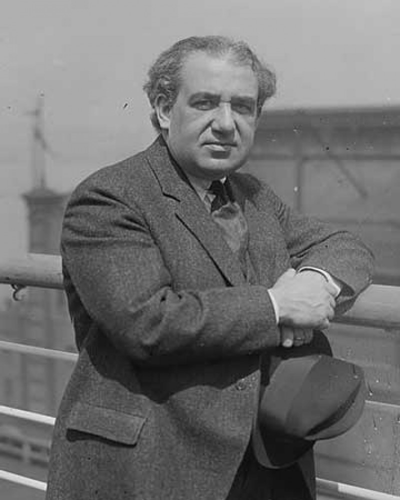 Man in a jacket and tie resting an elbow on a railing and holding a hat