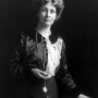 Woman holding a lorgnette and standing next to a table, with her hand on an open book