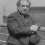 Man in a jacket and tie resting an elbow on a railing and holding a hat
